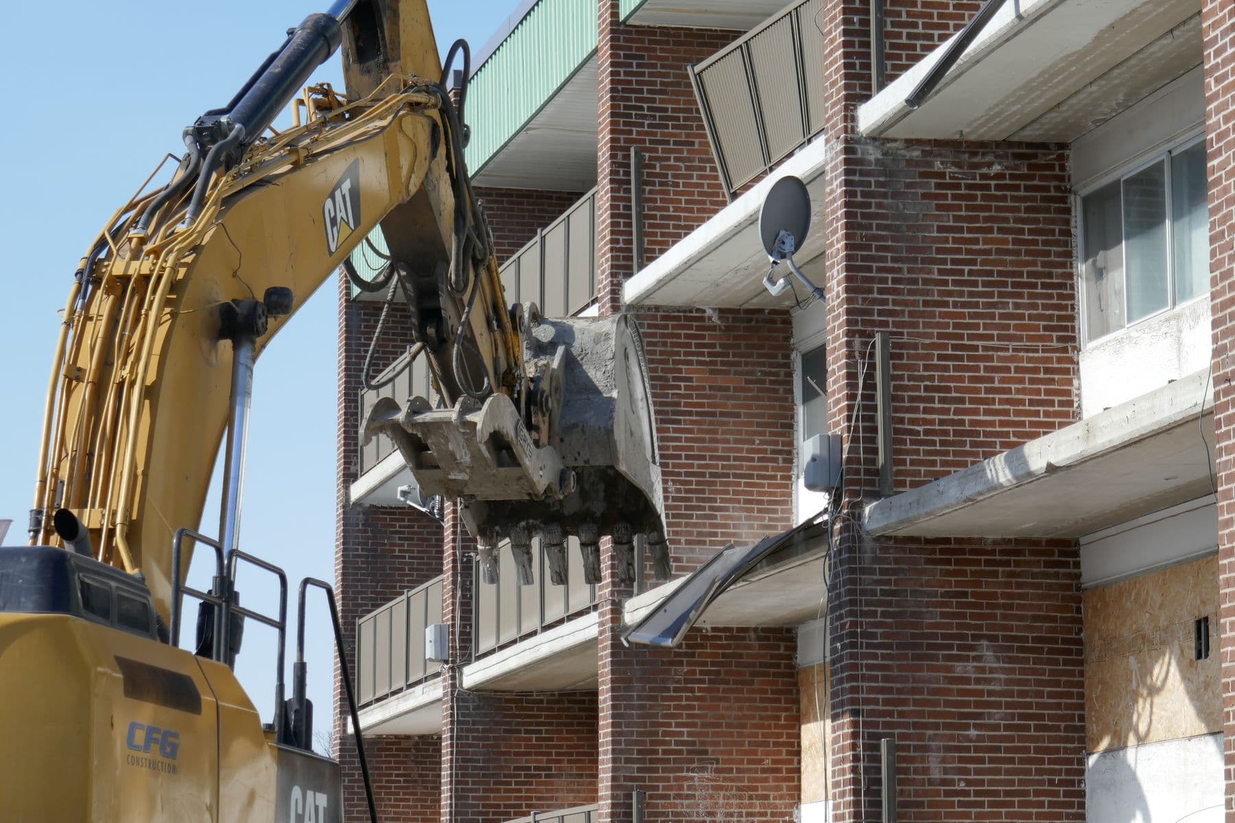 An excavator arm tearing the balconies off a three-story brick apartment building.