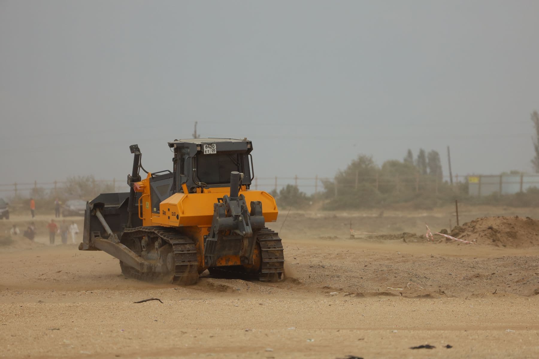 An orange bulldozer pushing across a bare, sandy lot under a gray sky.