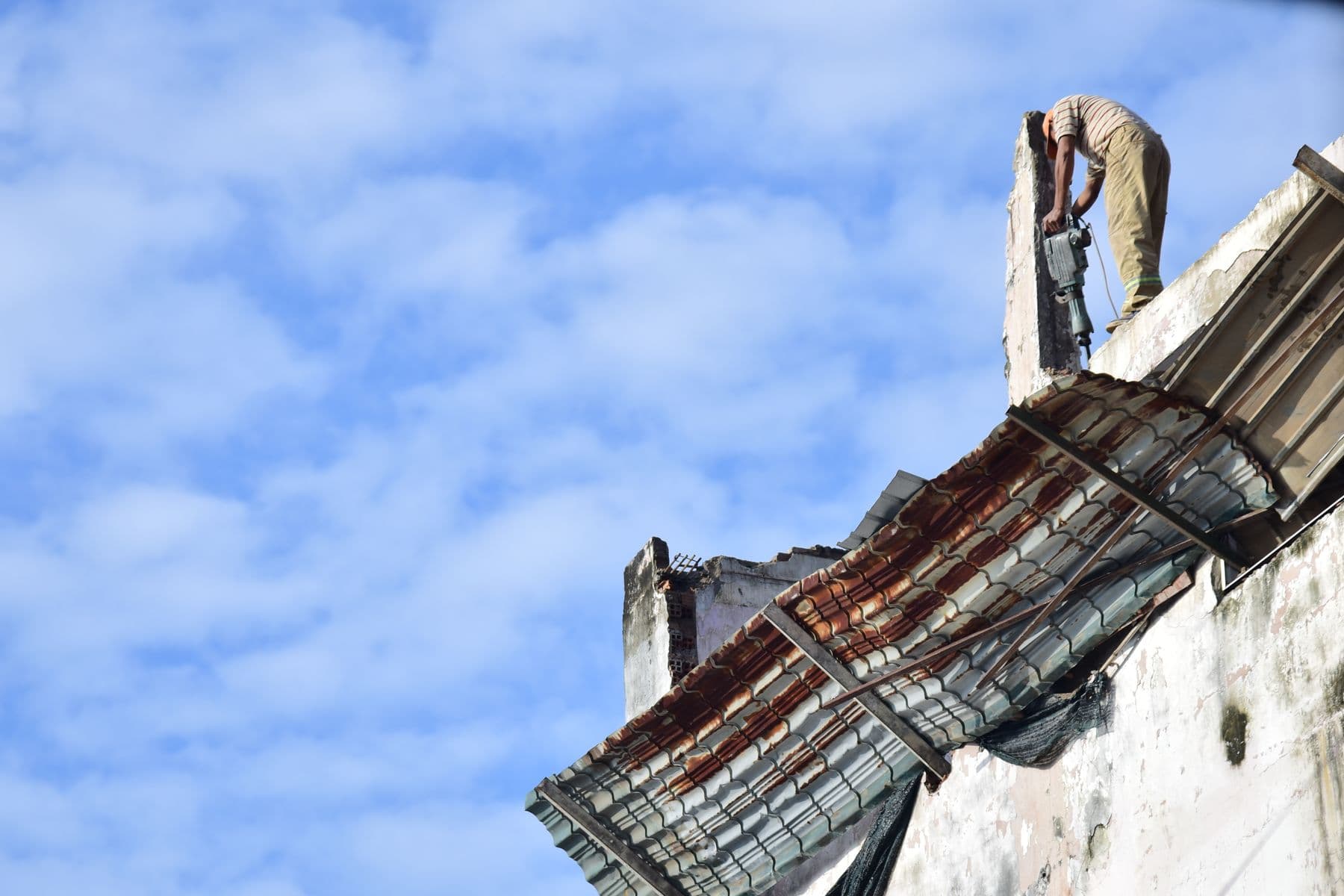 A worker standing on top of a wall, pulling a rusted corrugated metal roof panel off a building.