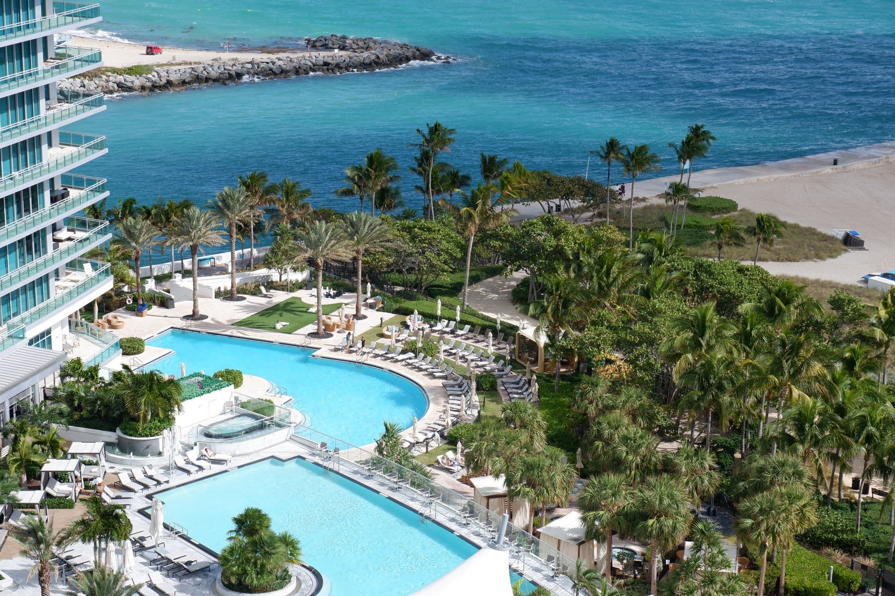 An aerial view of two hotel swimming pools among palm trees, between a glass tower and the beach.