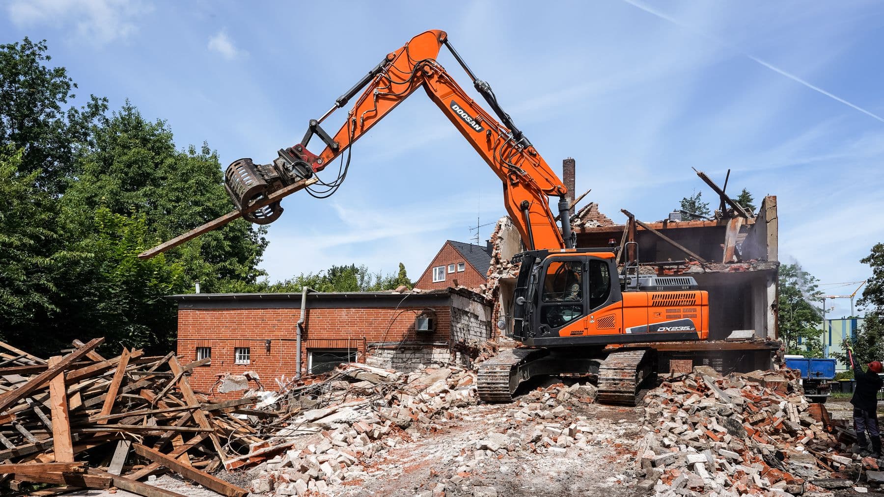 An orange excavator pulling apart a small brick building, with timber and broken brick piled in front.