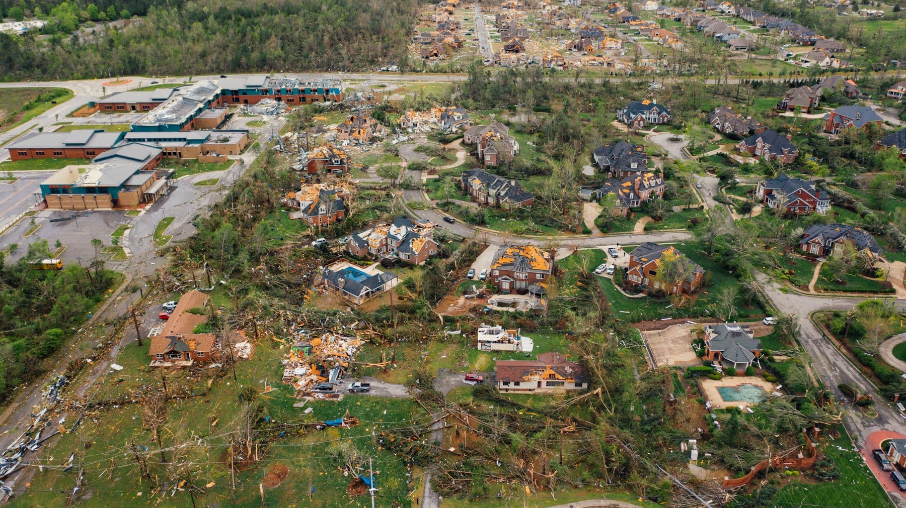 An aerial view of a neighborhood with damaged roofs and uprooted trees after a storm.